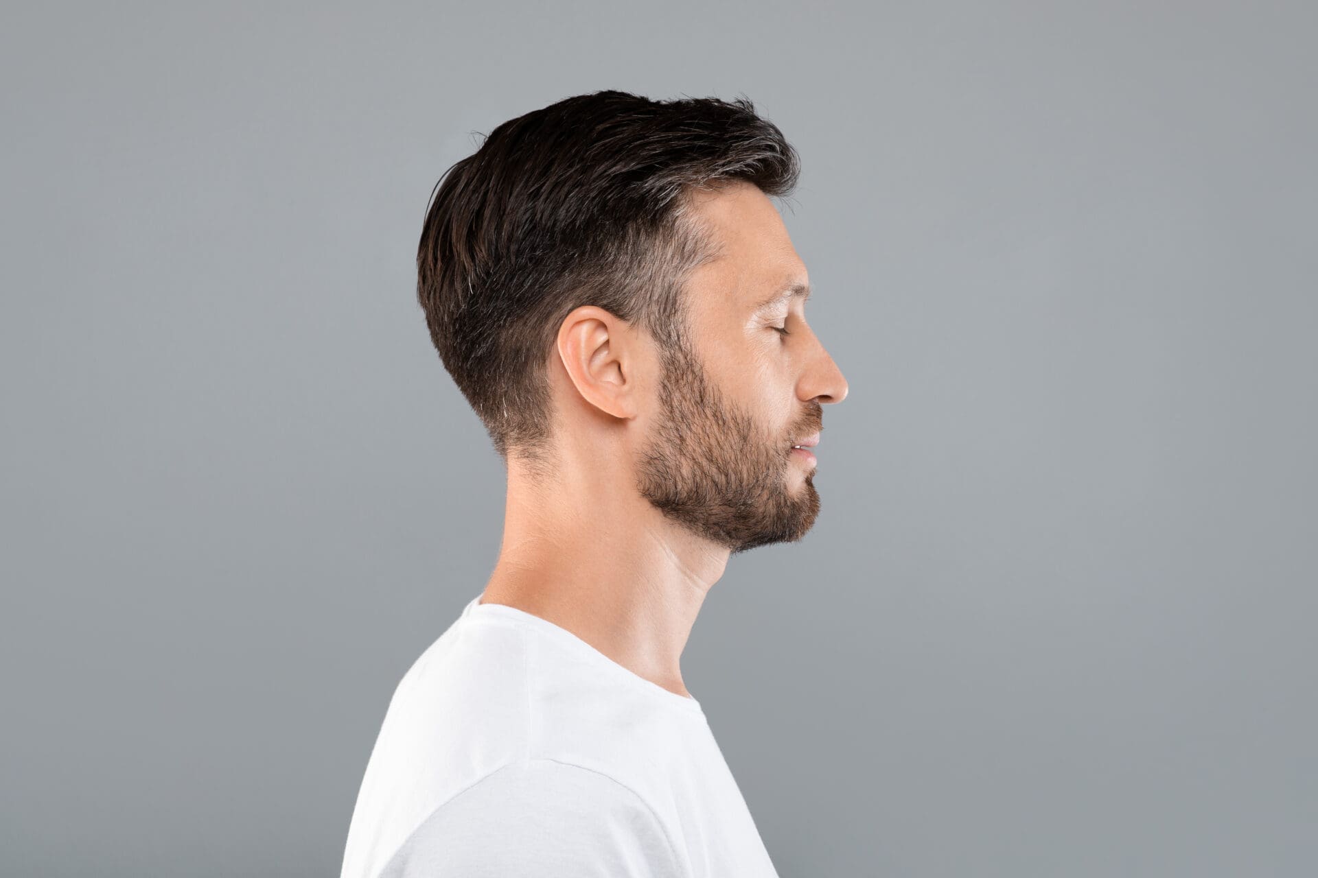 Side view of middle-aged man over grey background Side view of middle-aged bearded man in white t-shirt over grey studio background, copy space. Profile portrait of handsome man with closed eyes posing on gray, standing straight and looking aside